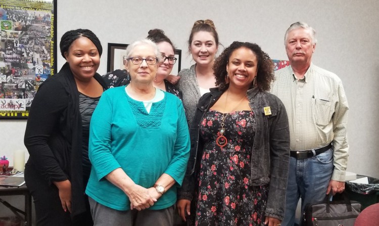 A group of 6 people that are intergenerational and racially diverse. There are 4 people in the back of the group. From left to right, a black woman smiles and is wearing a black sweater and grey patterned shirt, one white women wearing glasses and her dark brown hair in a bun smiles, another white woman smiles with her brown hair up in a bun, and a white older man in a button down shirt and jeans. In the front is an older woman with white hair and glasses. She's wearing a turquoise shirt and glasses. A brown skinned woman wears a flowered sun dress, jean jacket and gold jewelry with curly hair smiles.