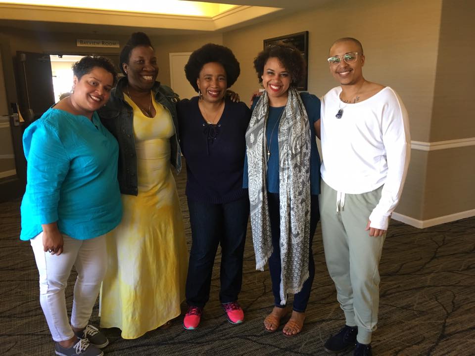 5 people of color stand with their arms around each others shoulders smiling at the camera.