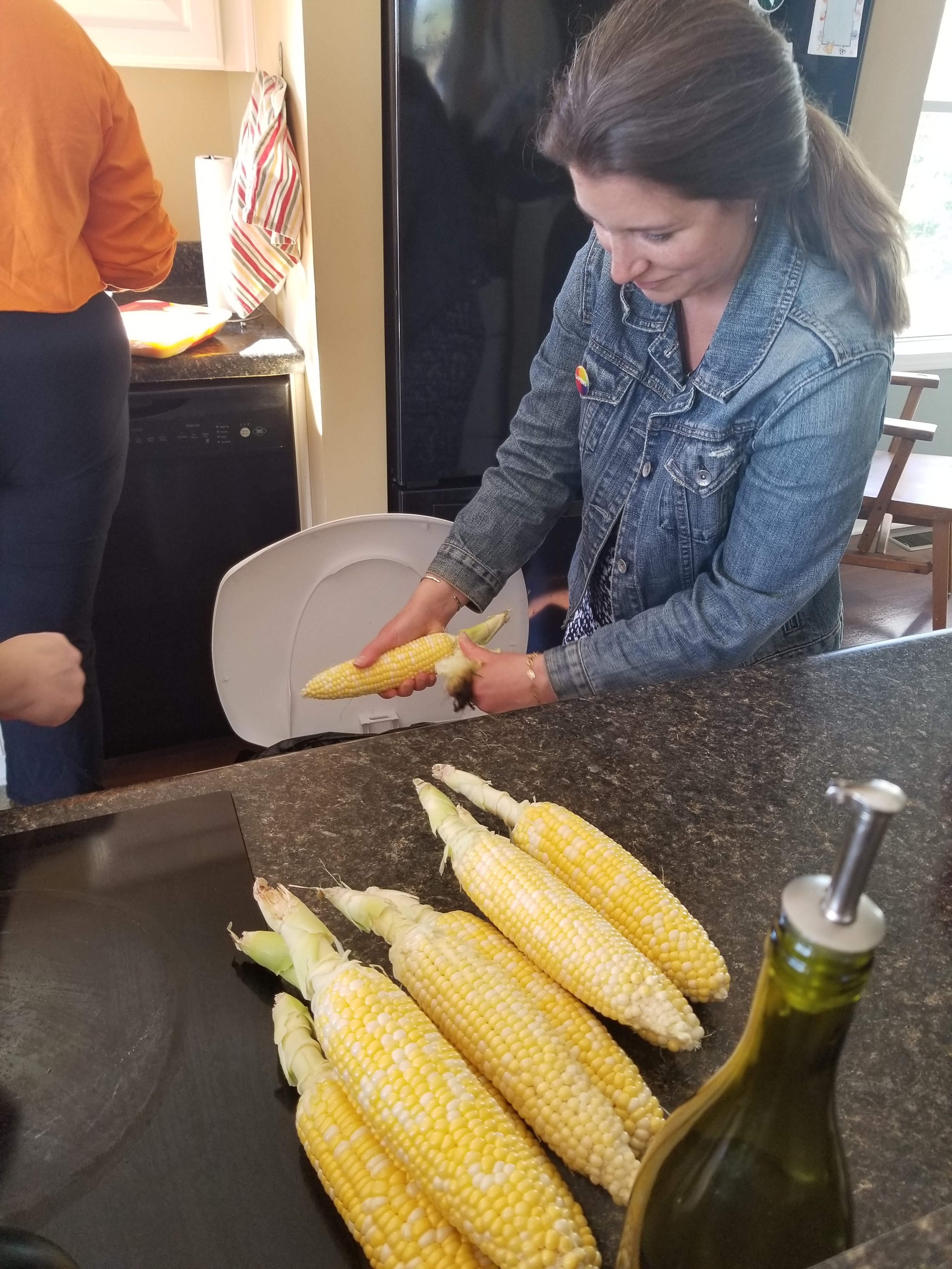 A white woman in a jean jacket shucks corn into a trash can. She is looking down and focused on the work. She is standing in a kitchen and shucked corn is in a pile on the counter in front of her. 