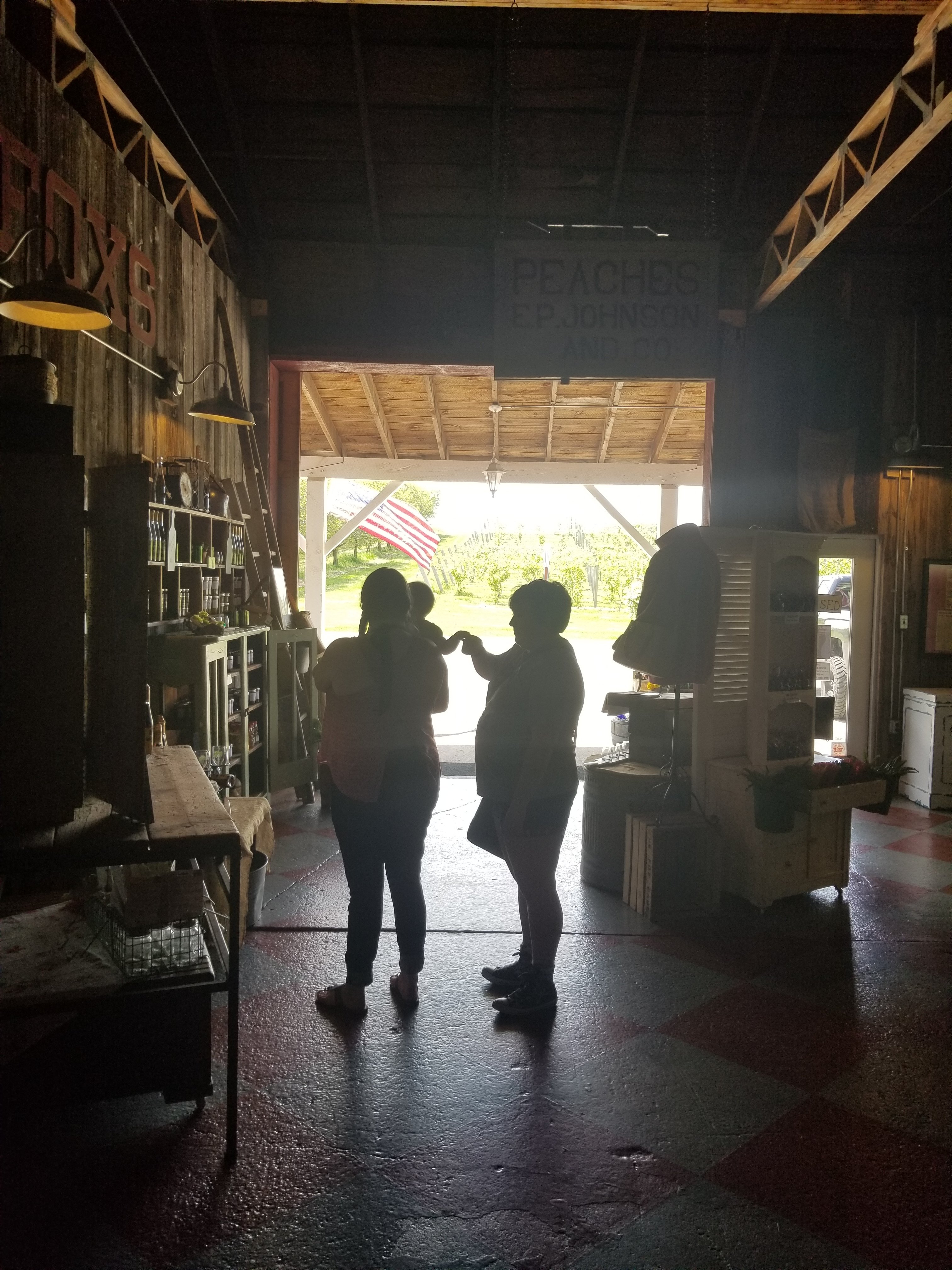 A person is holding a baby who is reaching out to another person who is touching the baby's hand. They are silhouettes in the open doorway of a barn marketplace. There are various shelves around them.